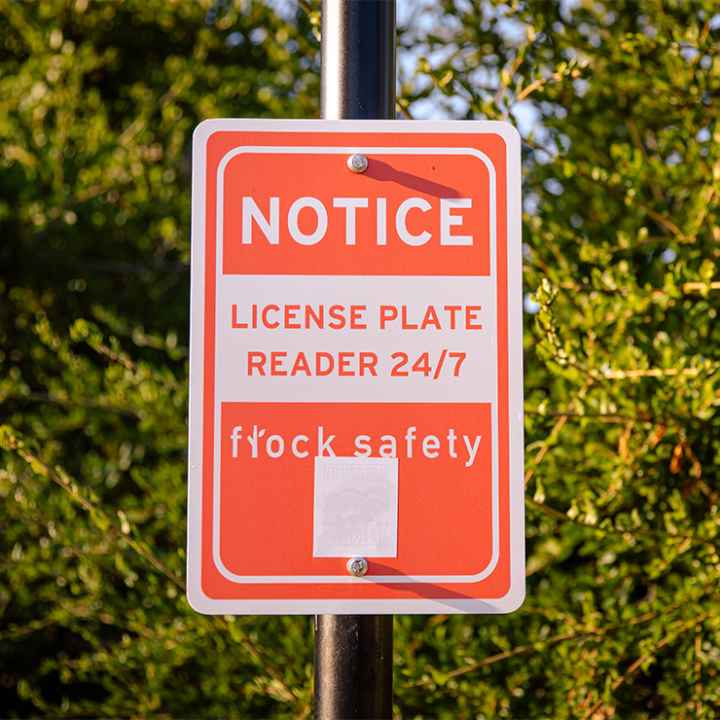 A sign attached to a metal pole with while letters on a red background reading "NOTICE _ LICENSE PLATE READER 24/7 - Flock Safety".
