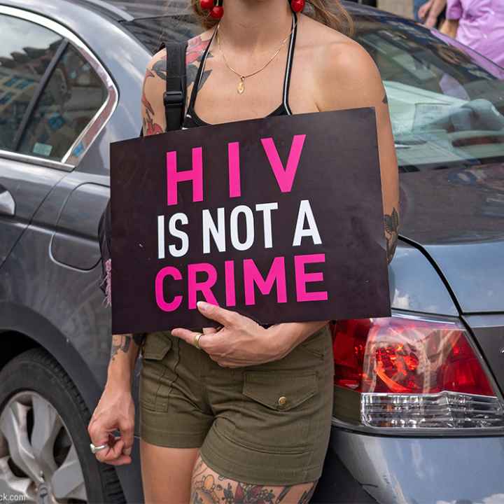A demonstrator, leaning against a light blue car and with their face hidden holds a sign reading "HIV IS NOT A CRIME".