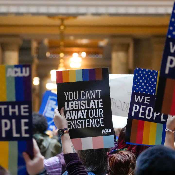 Individuals in a group holding ACLU-branded signs saying "We the People," and "You Can't Legislate Away Our Existence."